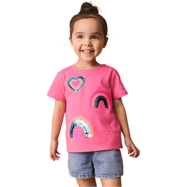 A toddler wearing a pink t-shirt with a rainbow and heart design, and denim shorts.